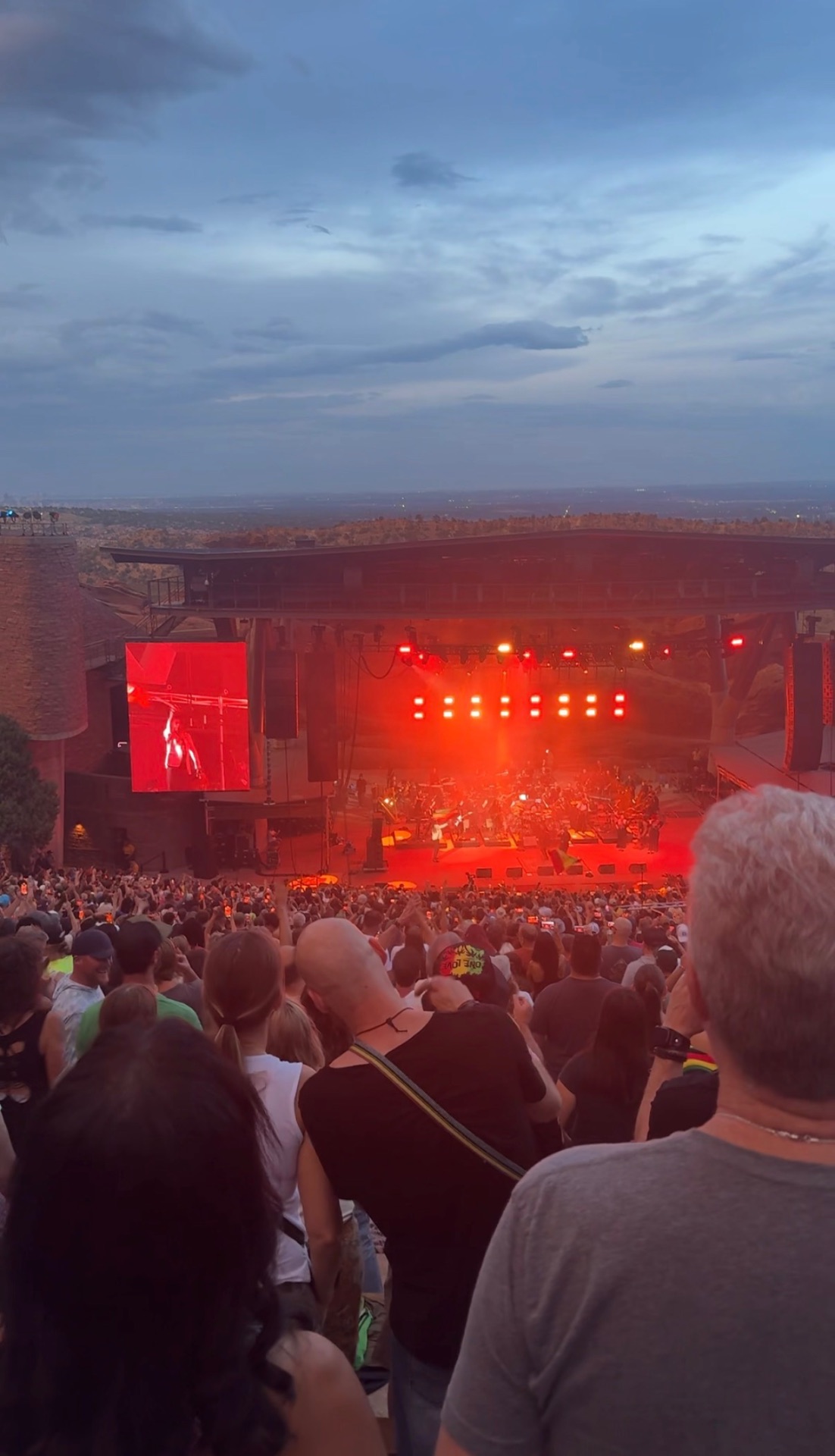 A packed concert at Red Rocks Amphitheatre near Morrison, Colorado, with the stage glowing red under a blue dusk sky