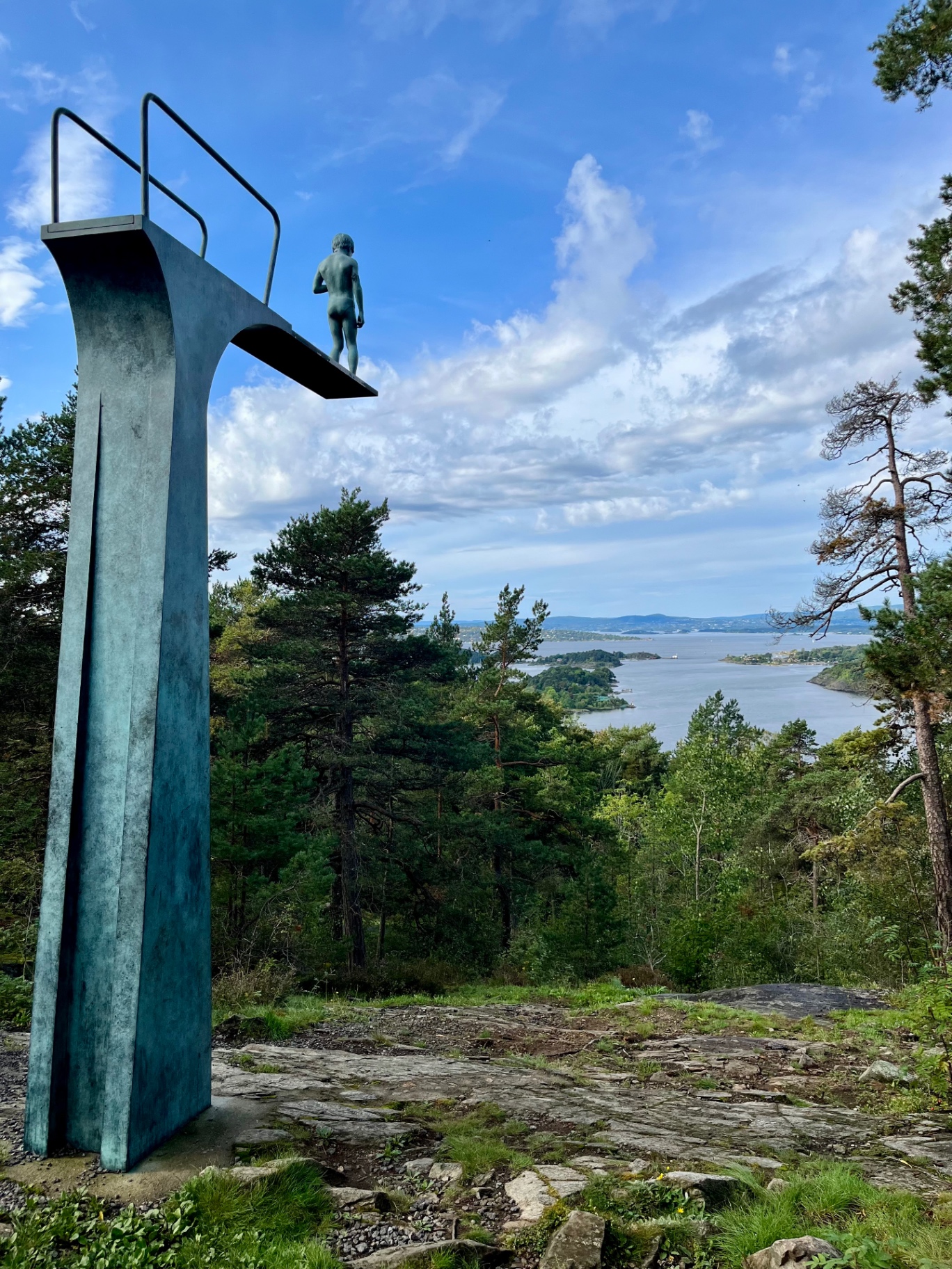 A bronze diving-board sculpture of a figure standing above the trees on a rocky headland, looking out over the Oslofjord in Norway.