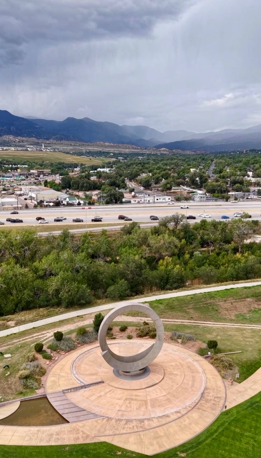 Aerial view of the Julie Penrose Fountain ring sculpture in America the Beautiful Park, Colorado Springs, with the highway, treeline and storm clouds over the Rocky Mountains beyond.