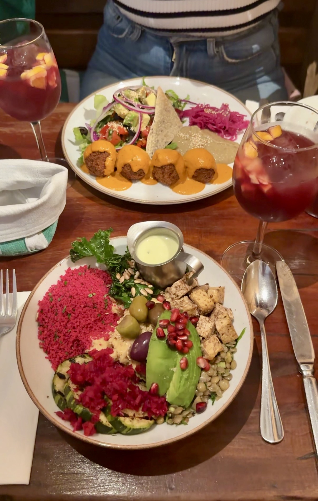 Two colourful vegetarian plates on a wooden table: a bowl with beetroot couscous, avocado, lentils, olives and tofu, and a plate of falafel with hummus, salad and pickled cabbage, beside glasses of sangria.