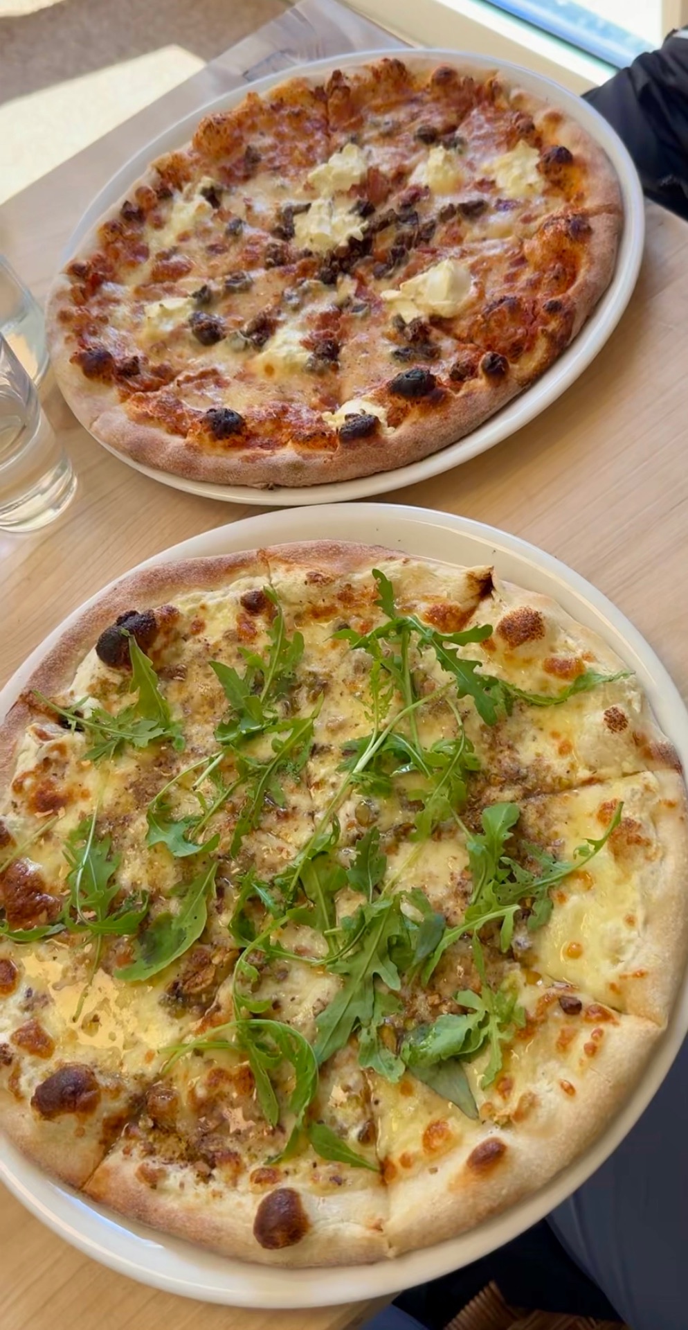 Two pizzas on white plates on a wooden table, the foreground pizza topped with melted cheese, crushed nuts and fresh arugula leaves