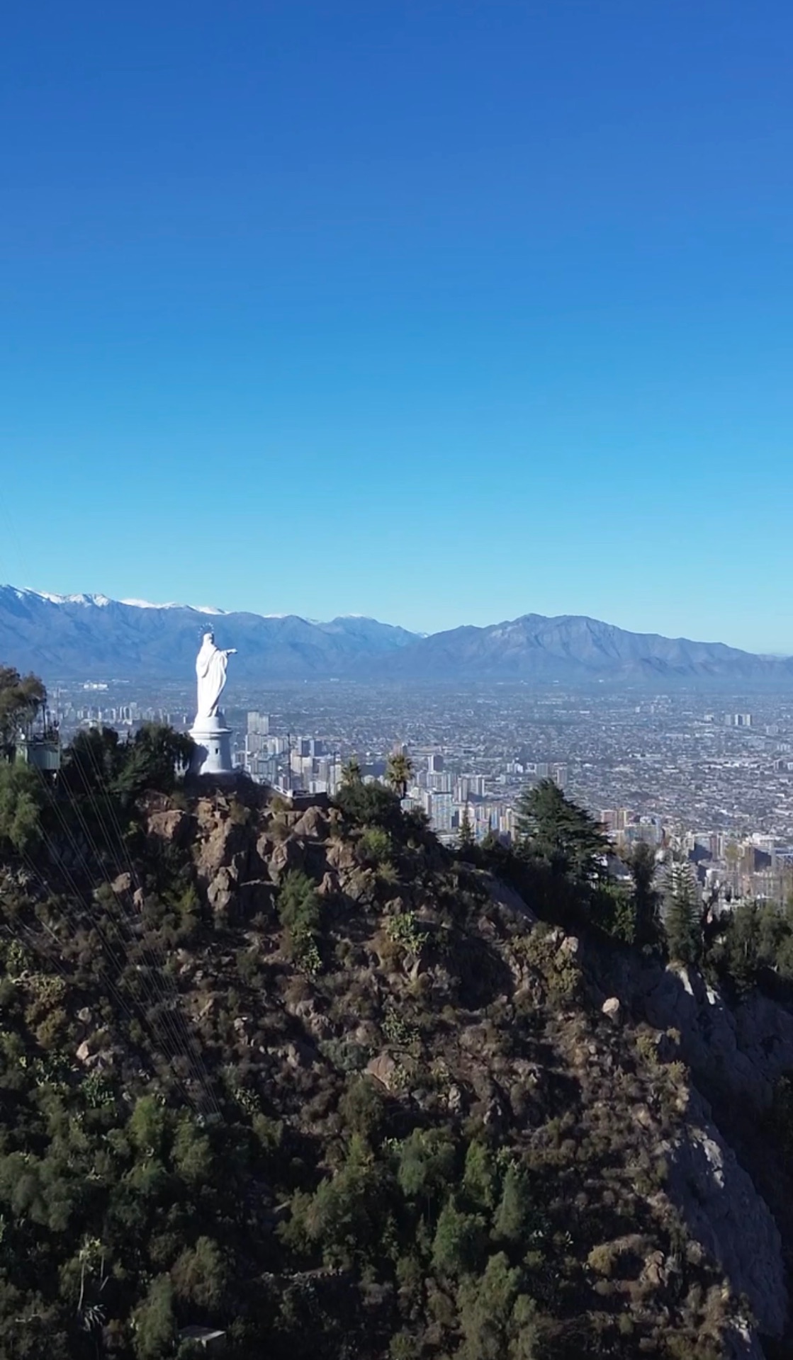 Aerial view of the white Virgin Mary statue on San Cristóbal Hill in Santiago, Chile, with the city spread out below and the snow-capped Andes on the horizon.