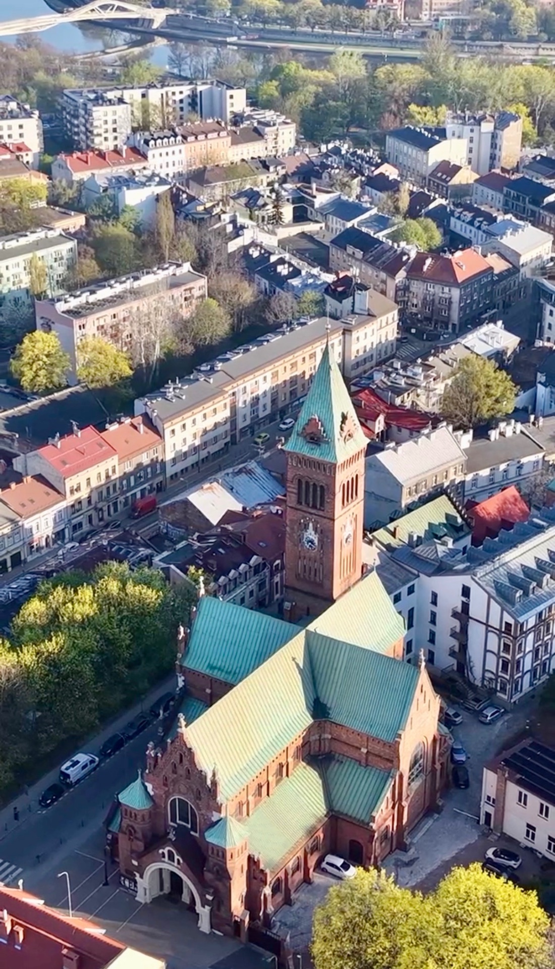 Aerial view of a red-brick church with a green copper roof and clock tower among Kraków rooftops, with the Vistula River beyond.