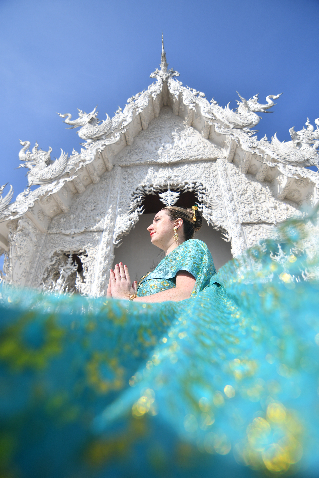 Miranda in traditional Thai clothing with her hands together in front of the ornate White Temple, turquoise fabric in the foreground