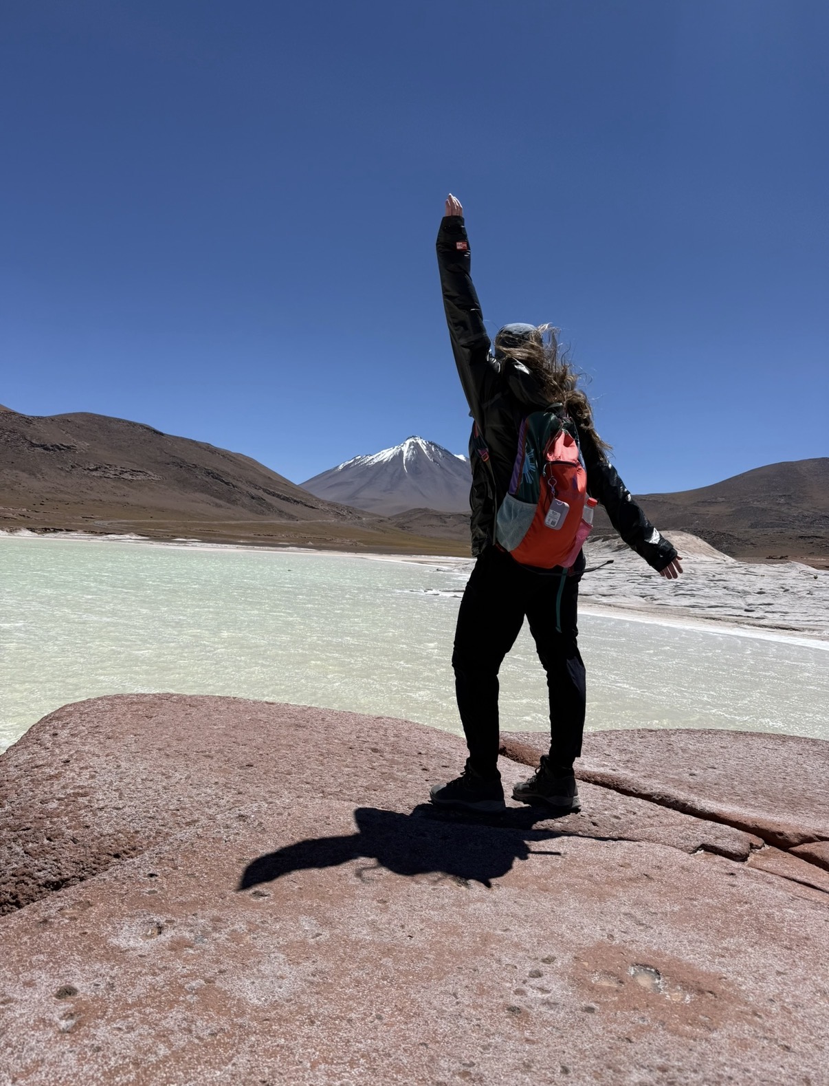 Miranda with one arm raised on red rocks beside a pale turquoise high-altitude lagoon at Piedras Rojas, with a snow-capped volcano behind.