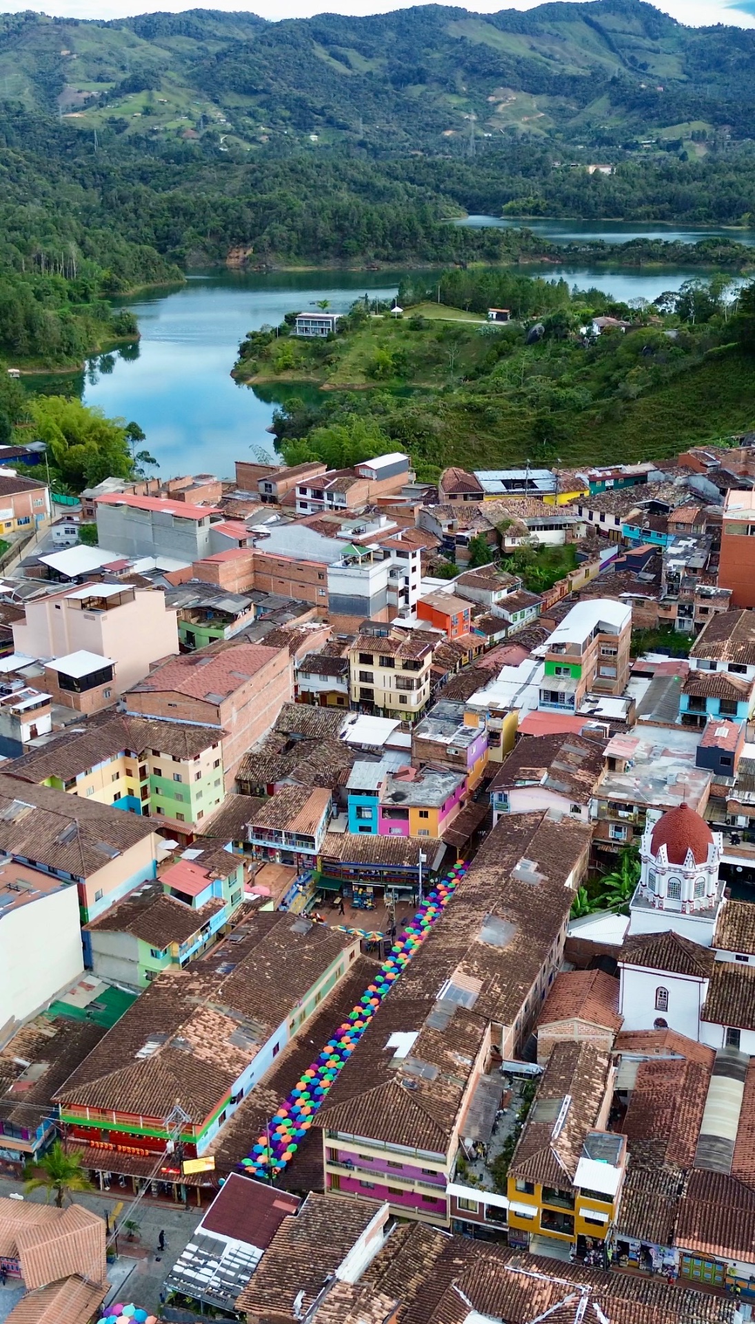 Aerial view of Guatapé, Colombia, looking along a street lined with rainbow umbrellas over painted zócalo houses and terracotta rooftops, with the reservoir and green hills behind.