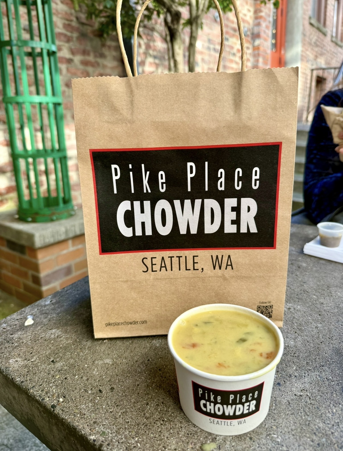A cup of vegan chowder from Pike Place Chowder in Seattle in front of its branded paper bag