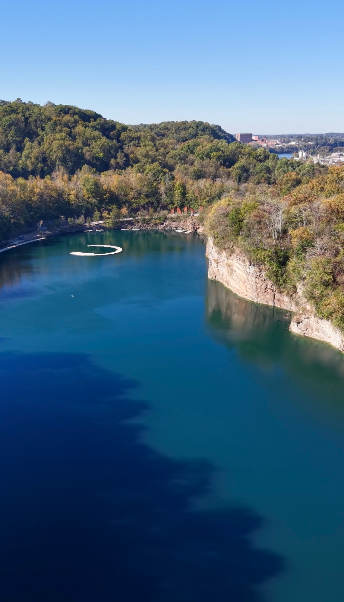 Aerial view of Fort Dickerson Quarry in Knoxville, with deep blue and emerald-green water, sheer tan rock cliffs and a curved white floating dock, forested hills and a brick building beyond