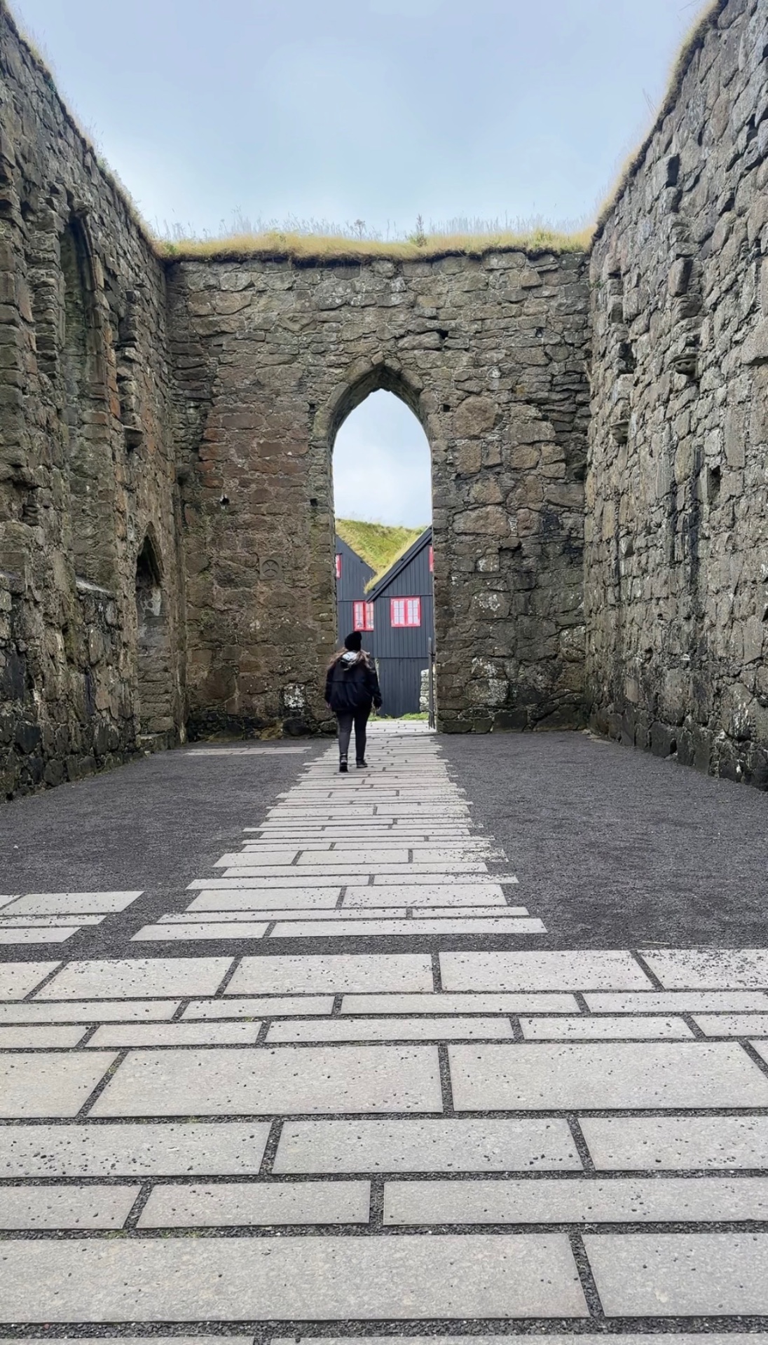 A visitor walking through the roofless stone ruins of Magnus Cathedral in Kirkjubøur, Faroe Islands, framing a black turf-roofed house with red windows beyond.