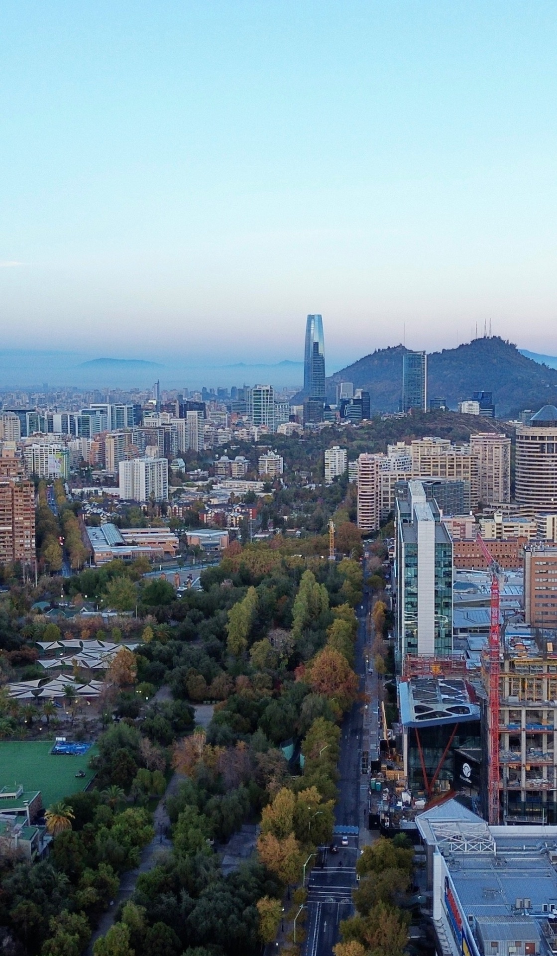 View from above over Parque Araucano and the Parque Arauco shopping district in Las Condes, Santiago, with high-rise buildings, a red construction crane, the Gran Torre Santiago and Cerro San Cristóbal on the horizon at dusk.