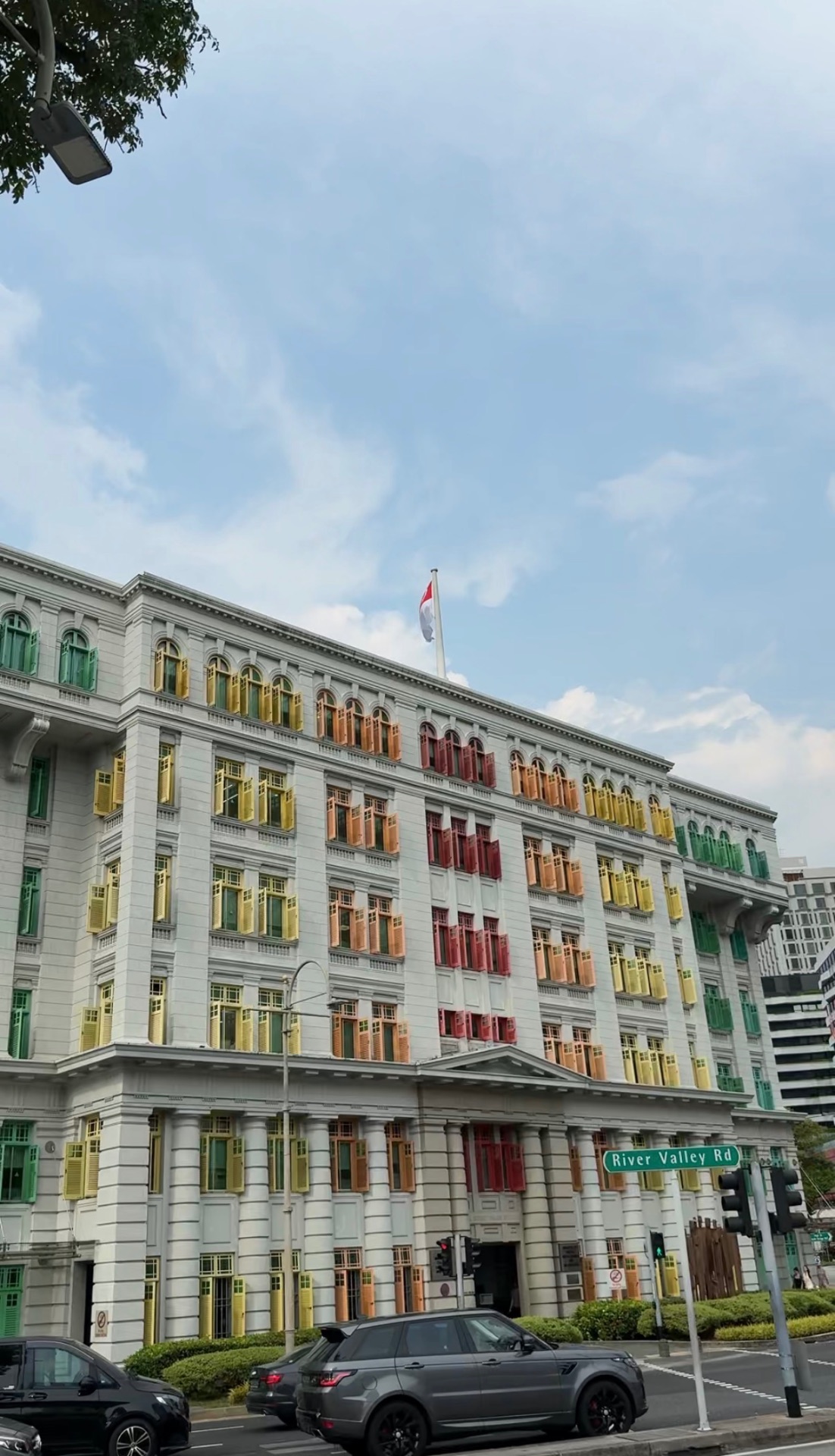 A grand neoclassical building in Singapore with colorful green, orange, red and yellow shuttered windows and a Singapore flag on the roof.