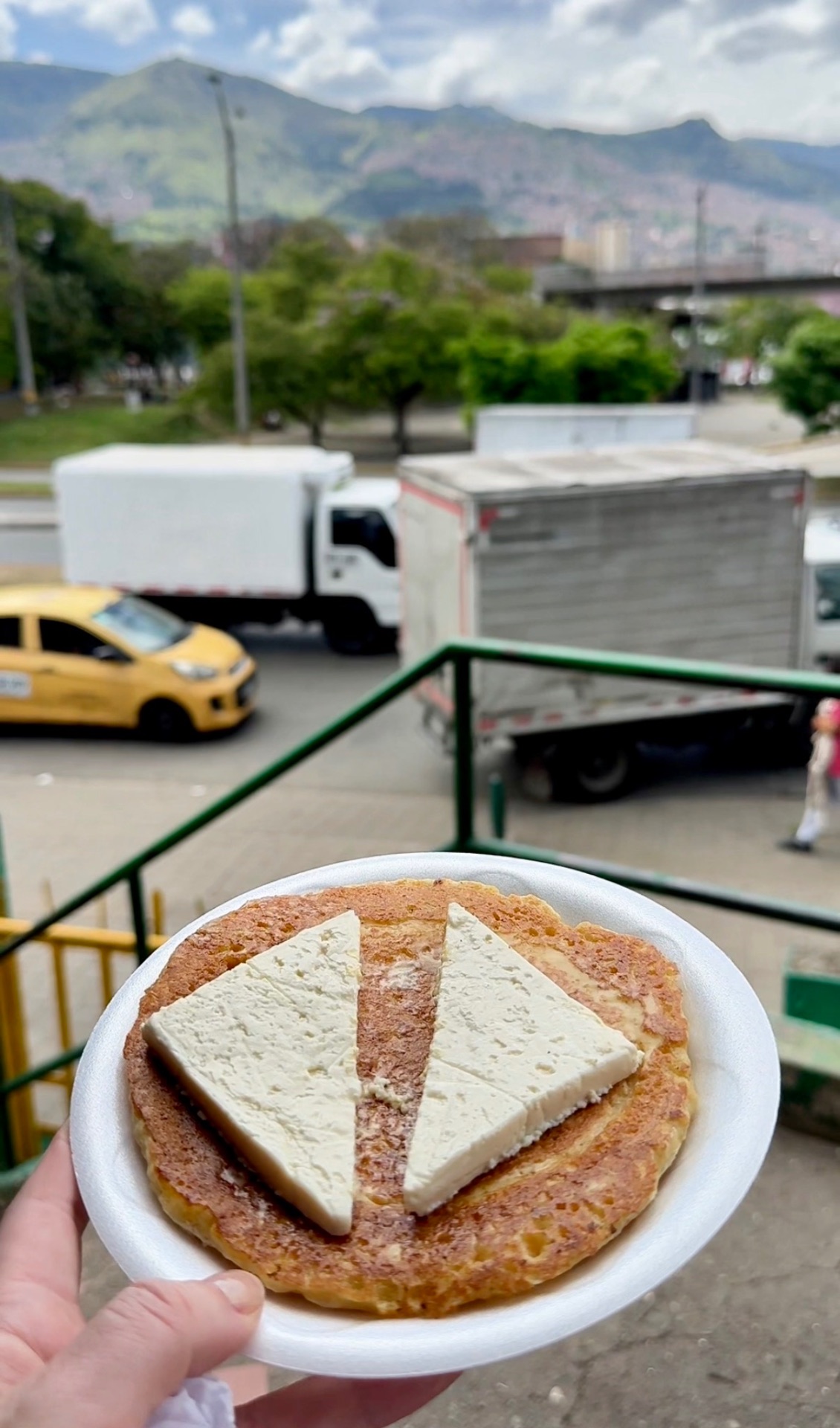 Freshly made arepa topped with cheese held up against the Medellín skyline at Plaza Minorista