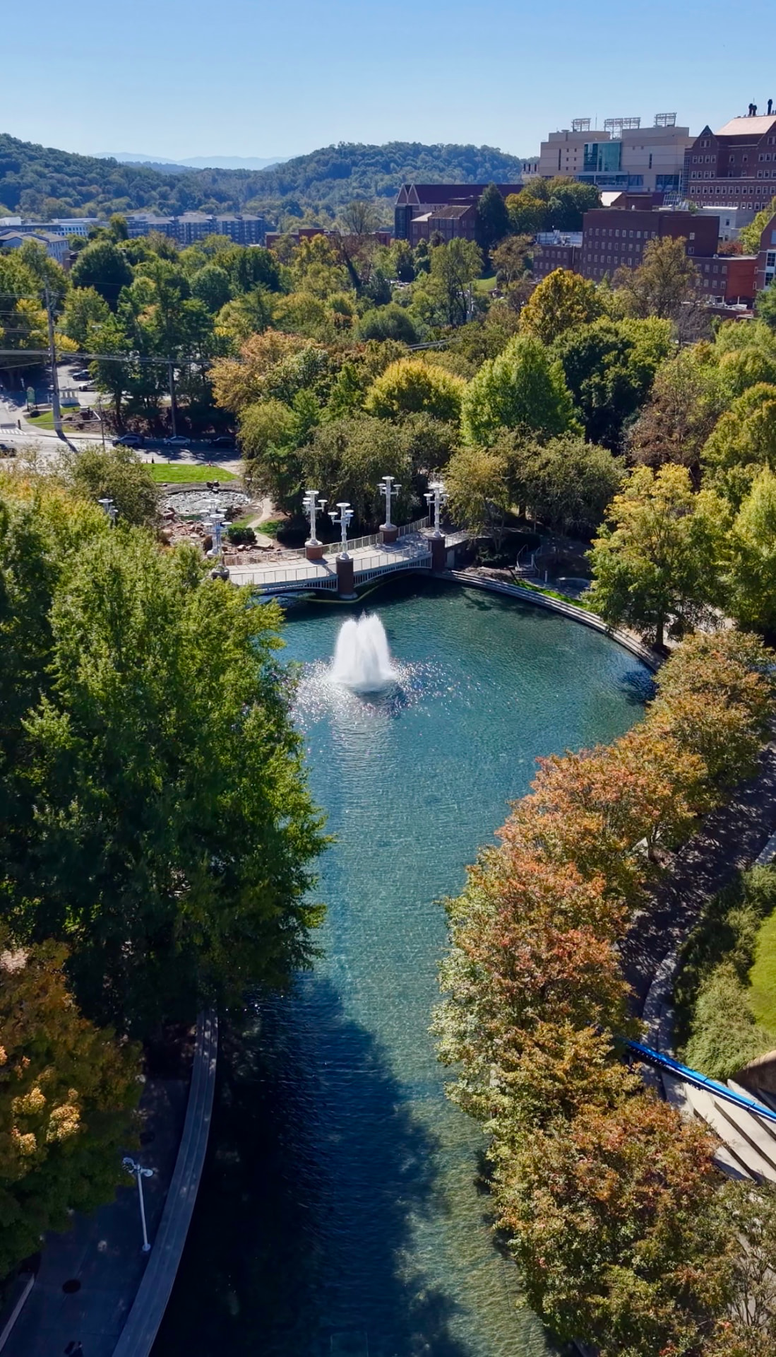 Drone view over World’s Fair Park in Knoxville, with a fountain in the turquoise lake, a footbridge and trees turning autumn colors