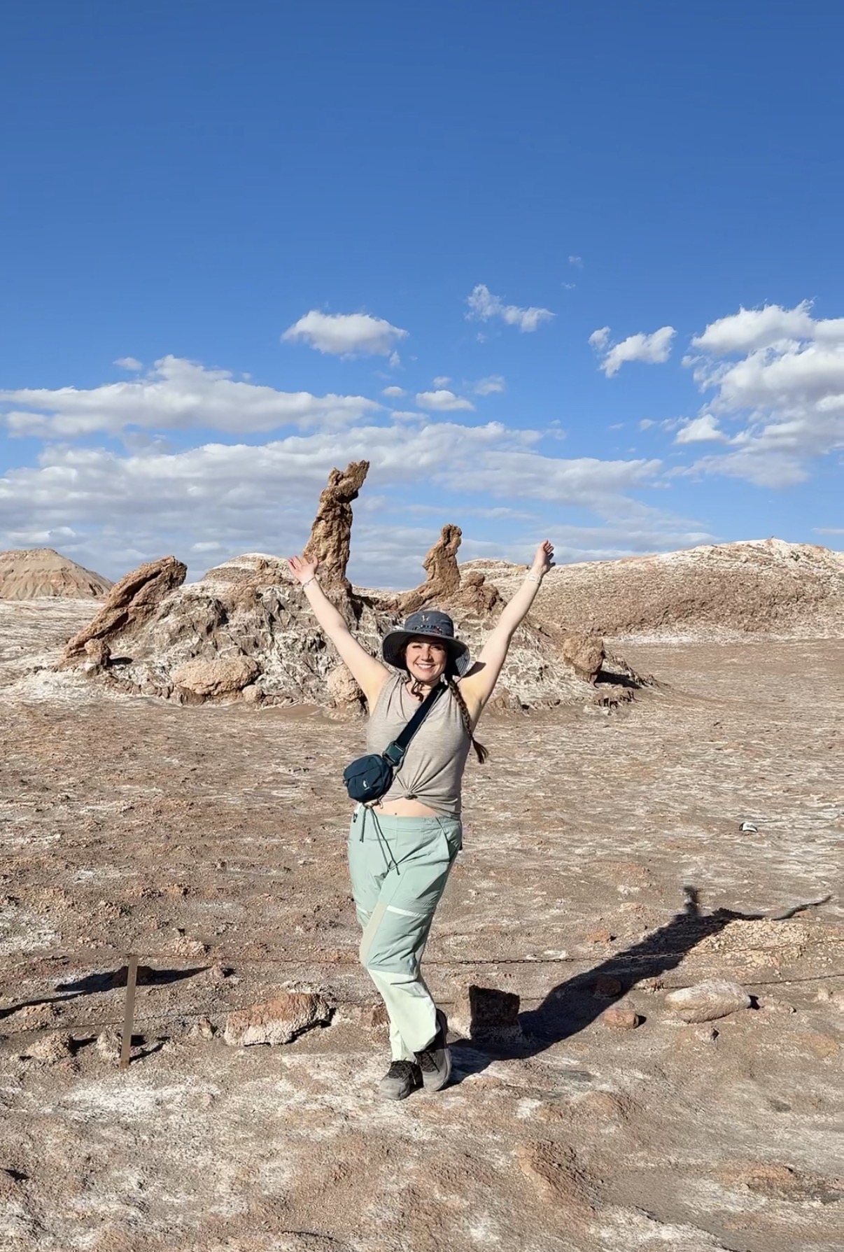 Miranda with arms raised in Valle de la Luna near San Pedro de Atacama, Chile, in front of the Tres Marías rock formations under a blue sky.
