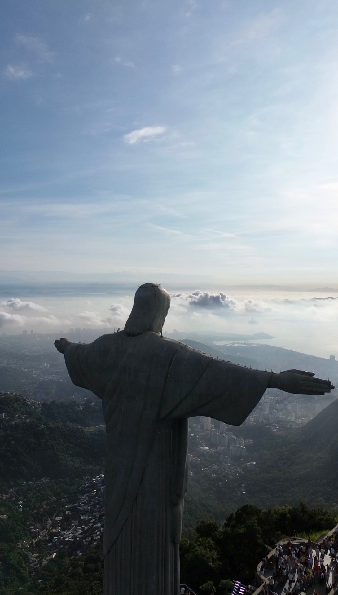 Aerial view from behind the Christ the Redeemer statue in Rio de Janeiro, looking out over the city, hills and ocean above a bank of cloud.